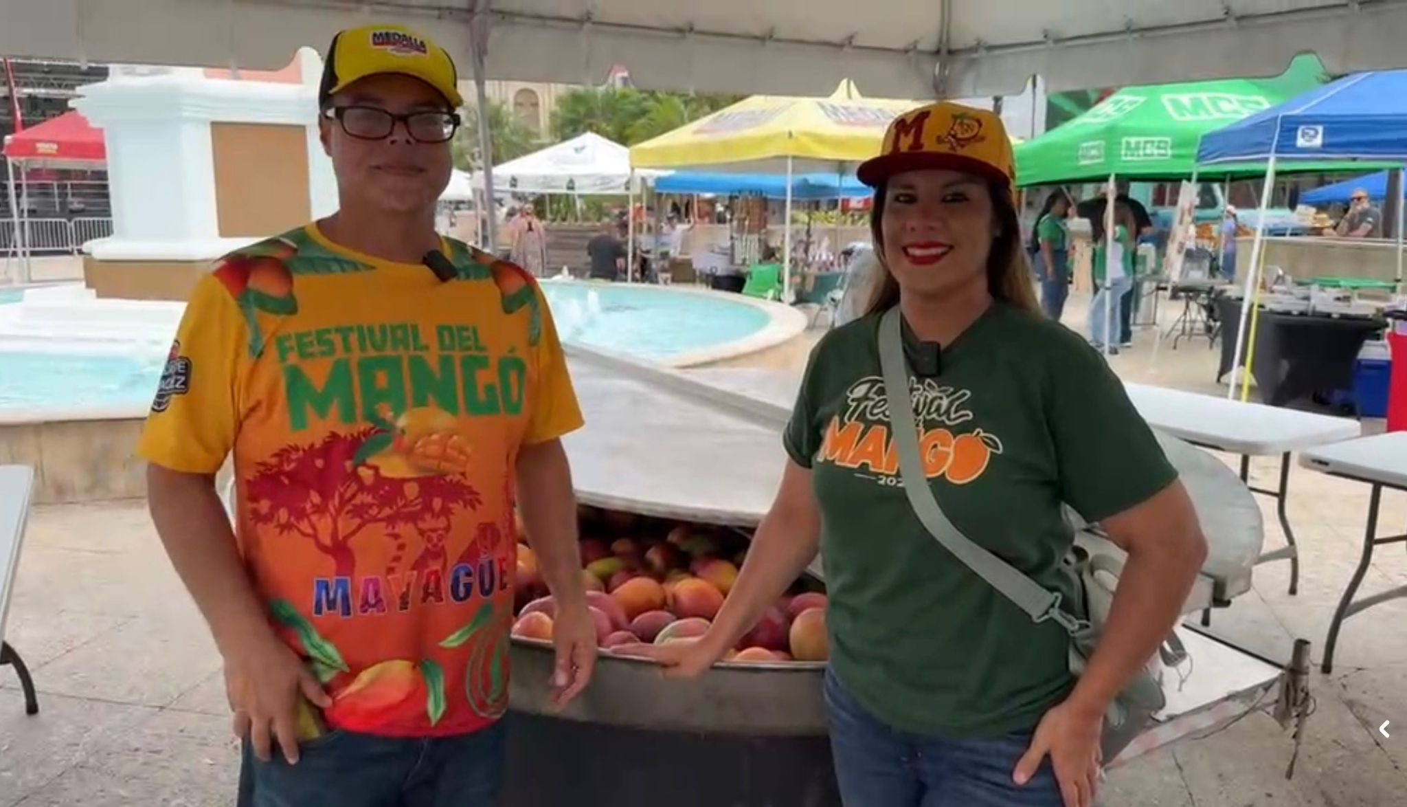 Presentadores del Festival del Mango con camisas oficiales anaranjada y verde frente a la fuente de la Plaza Pública